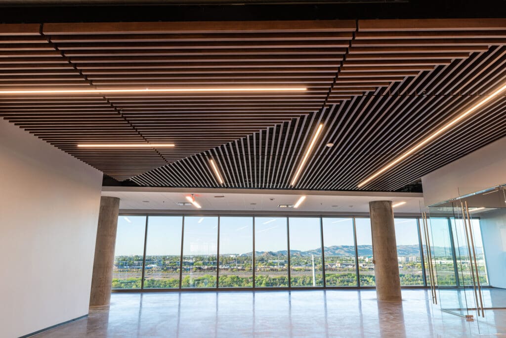 A modern, empty office space with a polished floor, a contemporary wood-slat ceiling with integrated lights, concrete pillars, glass walls, and large windows showcasing a scenic cityscape with green trees and mountains in the distance.
