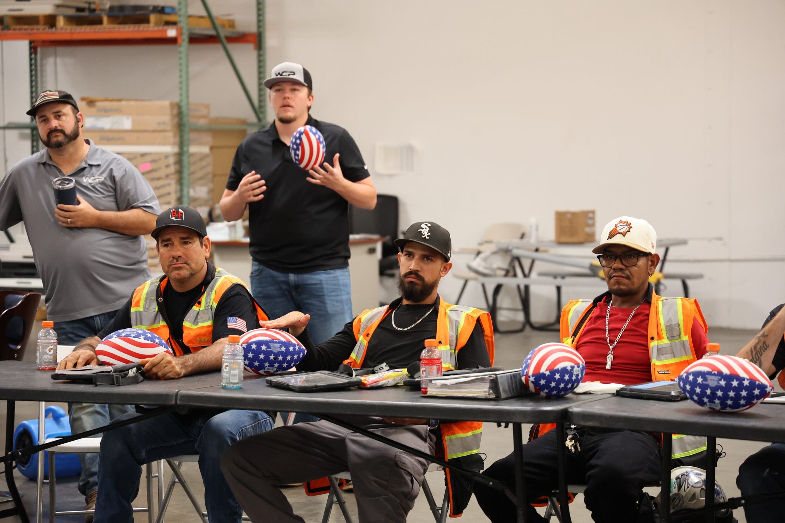 A group of four men wearing safety vests and hard hats with American flag designs sit at a table in a warehouse, listening intently as articles and important instructions are discussed. Another man stands behind them gesturing, with boxes and metal shelves in the background.