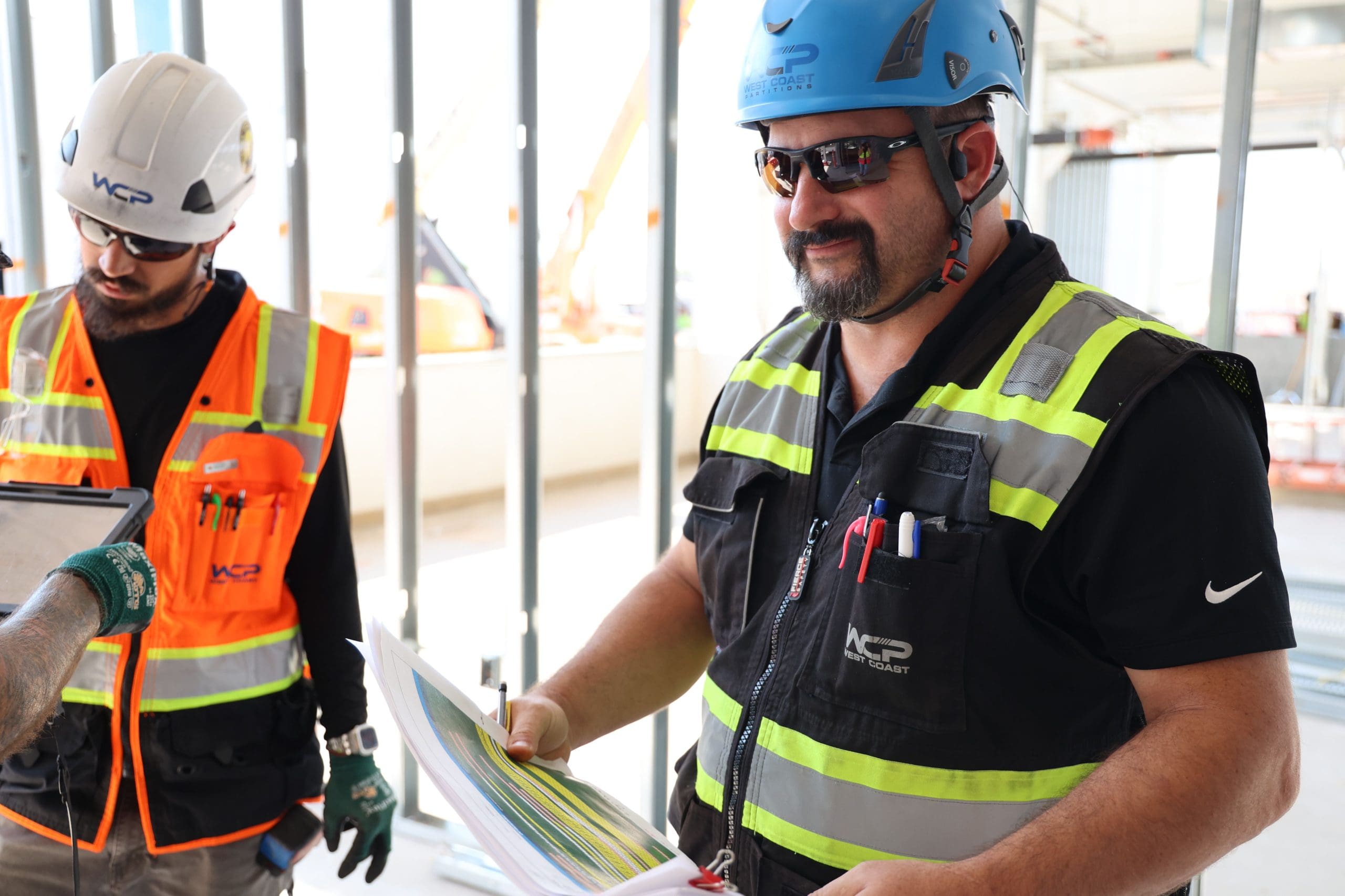 Two construction workers wearing safety vests and helmets stand indoors. One, holding articles and smiling, wears a blue helmet and sunglasses; the other, looking down at a tablet, wears a white helmet and orange vest. Metal framing is visible in the background.