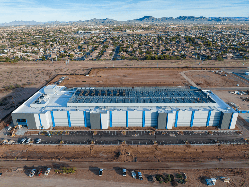 A large, modern data center with a white roof and blue vertical accents sits on a cleared dirt lot. Solar panels cover part of the roof. Articles and equipment are visible out front, alongside sparse landscaping and parked vehicles, with homes and mountains beyond.