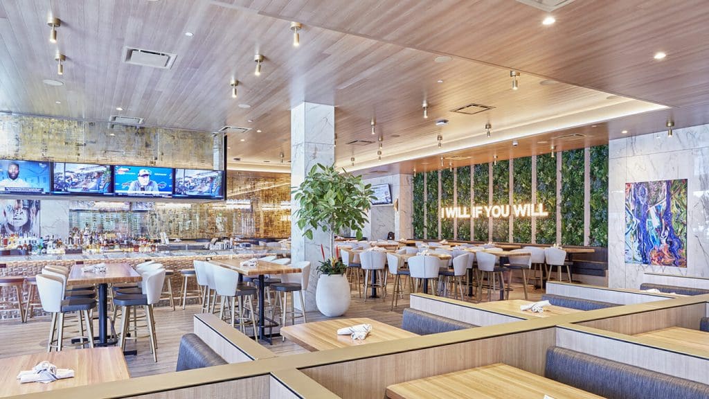 Modern, bright restaurant interior with wood and marble decor, West Coast Partitions, green plant wall featuring a neon "WILL IF YOU WILL" sign, multiple TVs above a bar, high tables, cozy booths with napkins, and a potted tree for a stylish welcome.