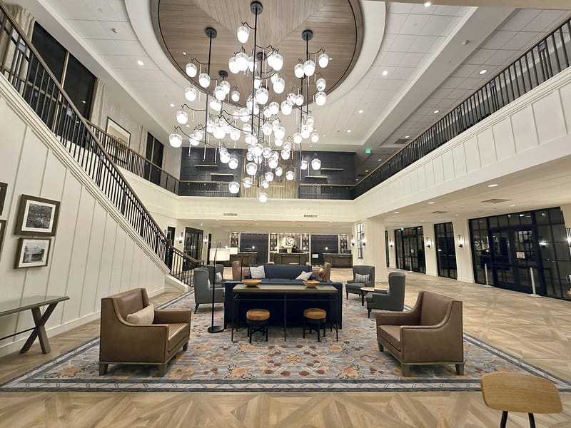 Spacious hotel lobby with modern chandelier, high ceiling, and open second-floor balcony. West Coast Partitions frame the seating area, which features tan leather chairs, gray armchairs, a blue sofa, and a patterned rug on chevron wood floors.