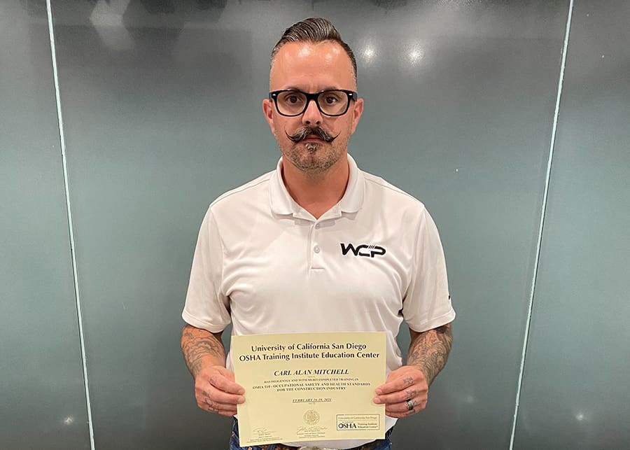 A man with glasses and a handlebar mustache, wearing a white collared shirt with a "WCP" logo, stands against a plain, frosted glass background, holding up a certificate from the University of California San Diego OSHA Training Institute Education Center.