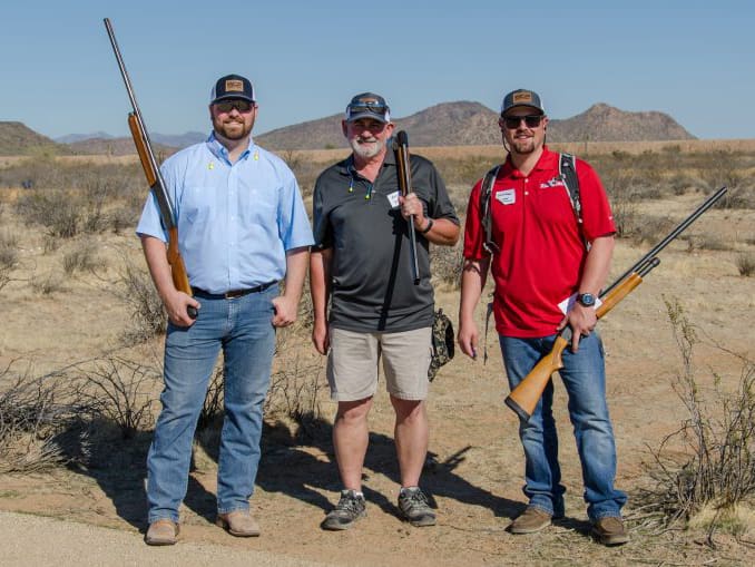 Three men stand outdoors in a dry, grassy landscape with mountains in the background. Each holds a shotgun. They wear sunglasses, caps, and casual clothes—one in a red shirt, one in a black shirt, and one in a blue shirt. All are smiling.