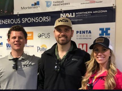 Three smiling people, two men and one woman, stand together in front of a sponsor banner. They all wear WCP-branded hats and shirts. The woman has long blonde hair and wears sunglasses and a pink shirt, while the men wear gray shirts.