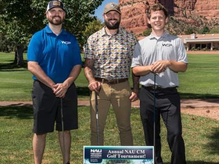 Three smiling men stand on a green golf course with red rock cliffs in the background. Each holds a golf club and wears a golf shirt and cap. In front of them is a sign reading “Annual NAU CM Golf Tournament!” with NAU and sponsor logos.