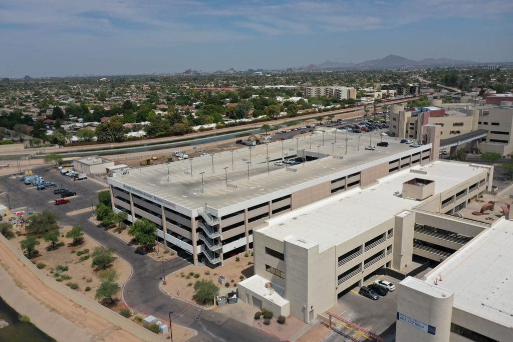 An aerial view of a multi-level parking garage surrounded by desert landscaping, nearby buildings, and a canal. Cars are parked on the roof and around the structure. In the background, there’s a cityscape with scattered homes, trees, and mountains under a blue sky.