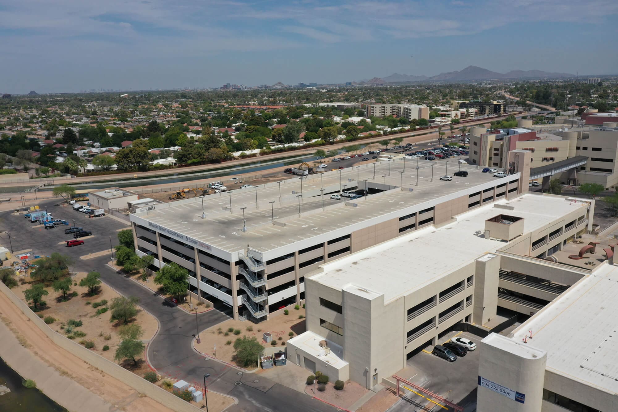 An aerial view of a multi-level parking garage surrounded by desert landscaping, nearby buildings, and a canal. Cars are parked on the roof and around the structure. In the background, there’s a cityscape with scattered homes, trees, and mountains under a blue sky.