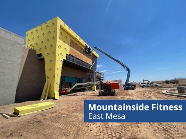 A construction site for a new Mountainside Fitness building in East Mesa. The structure is partially covered with yellow insulation panels. A crane lifts workers to the upper section. Various construction vehicles are on the dirt lot under a clear blue sky.