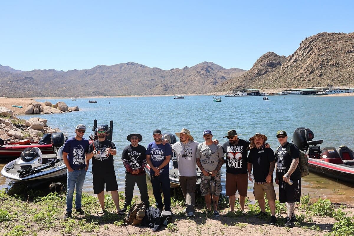 Ten men stand in a row on a grassy lake shore with fishing boats in the water behind them. They wear casual clothes, hats, and sunglasses. Rocky hills and clear blue sky are in the background, with more boats and a dock visible across the lake.
