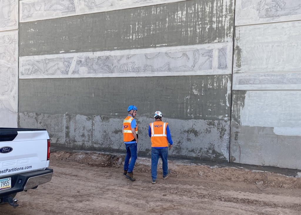 Two construction workers in orange safety vests and hard hats stand near a large concrete wall with patches of wet cement. A white pickup truck is parked nearby on the dirt ground. The workers appear to be inspecting or discussing the wall.