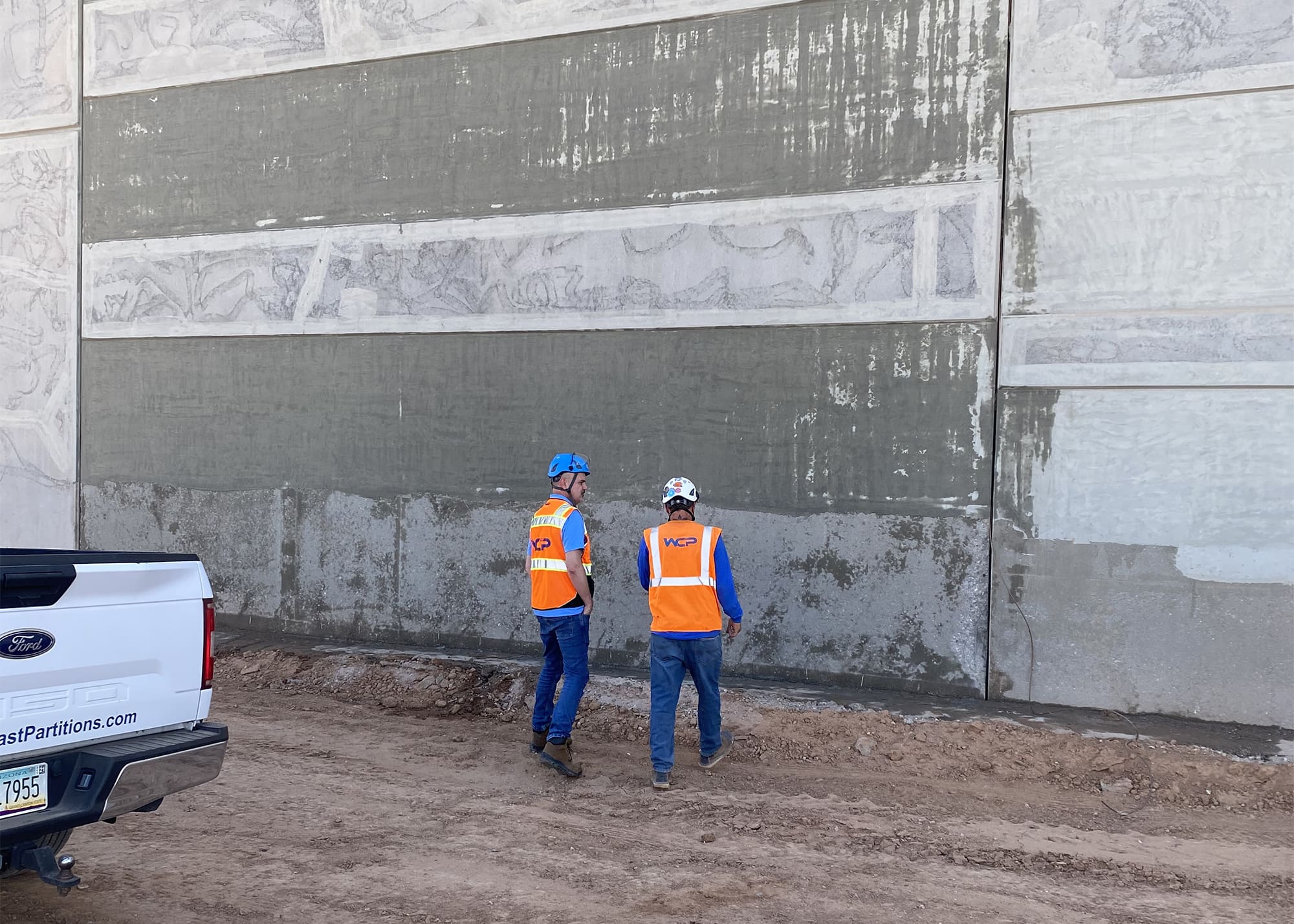 Two construction workers in orange safety vests and hard hats stand near a large concrete wall with patches of wet cement. A white pickup truck is parked nearby on the dirt ground. The workers appear to be inspecting or discussing the wall.