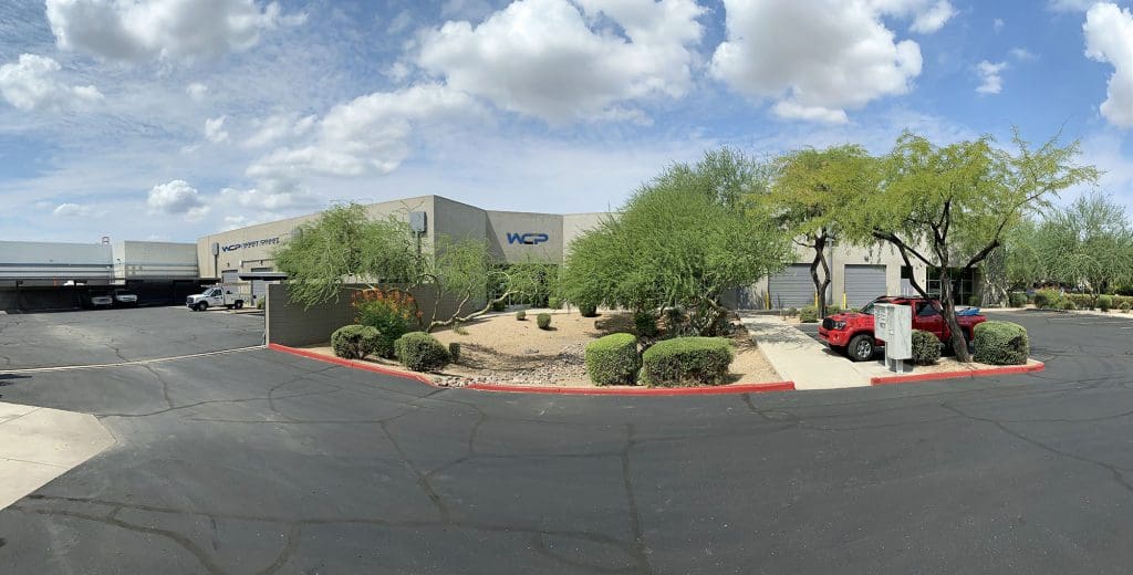 A modern industrial building with "WCP" on the facade, surrounded by desert landscaping with small trees and shrubs. A red vehicle is parked by the entrance. The sky is partly cloudy, and the parking lot appears empty.