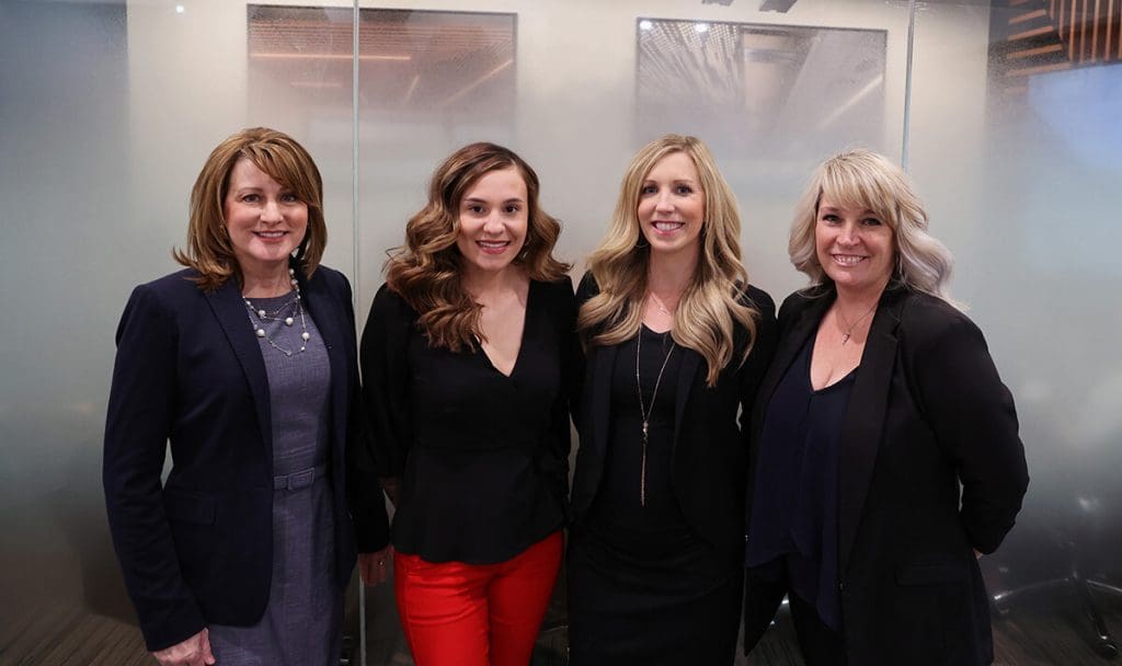 Four women stand together indoors, smiling at the camera. They are dressed in business attire: three wear dark blazers, and one wears a black top with bright red pants. The background features frosted glass panels and a modern office setting.