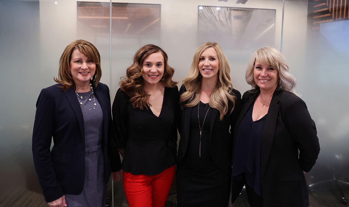 Four women stand together indoors, smiling at the camera. They are dressed in business attire: three wear dark blazers, and one wears a black top with bright red pants. The background features frosted glass panels and a modern office setting.