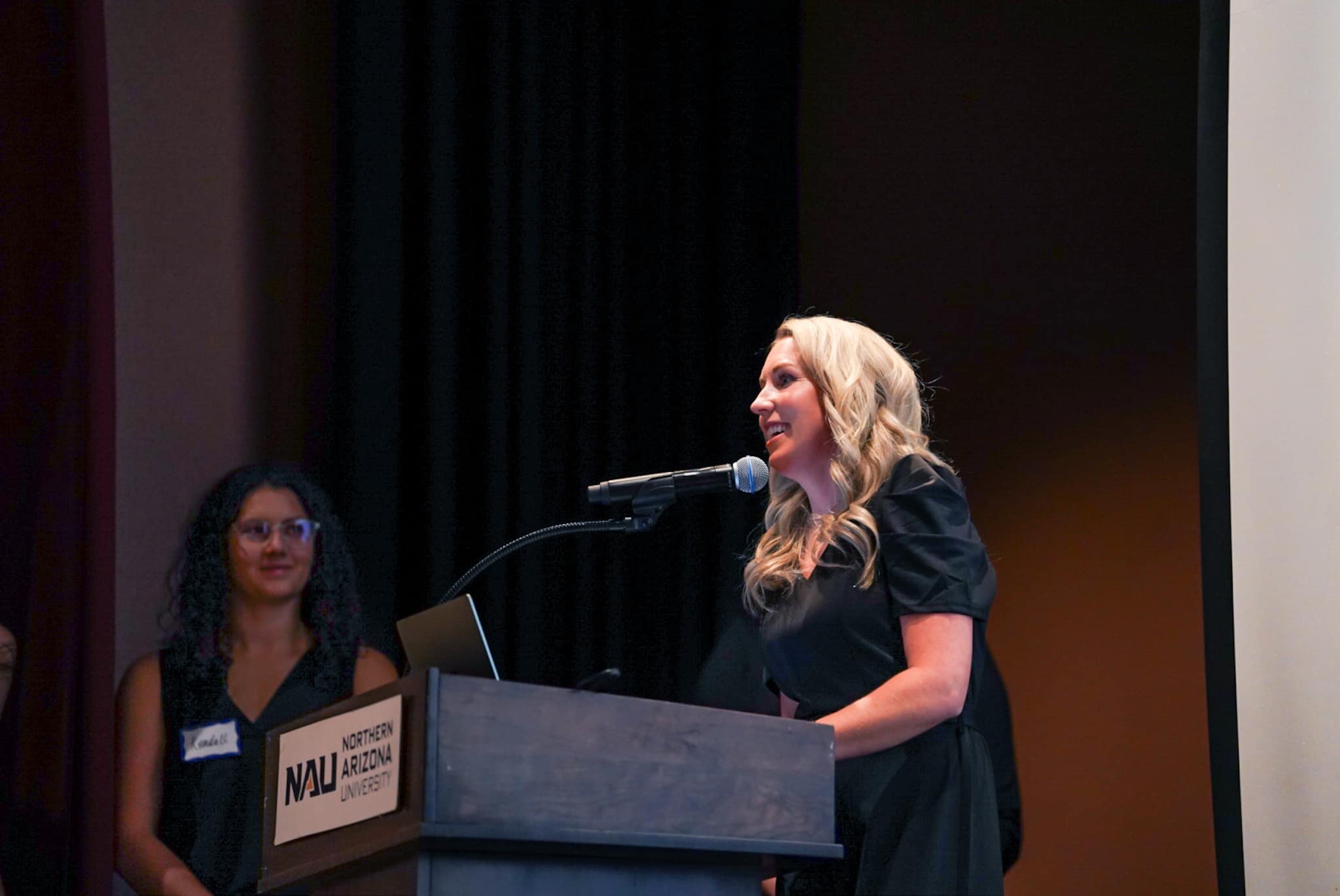 A woman with long blonde hair in a black dress speaks at a podium labeled “Northern Arizona University.” A microphone is attached to the podium. Another woman with curly hair and glasses stands in the background, partially visible and smiling.