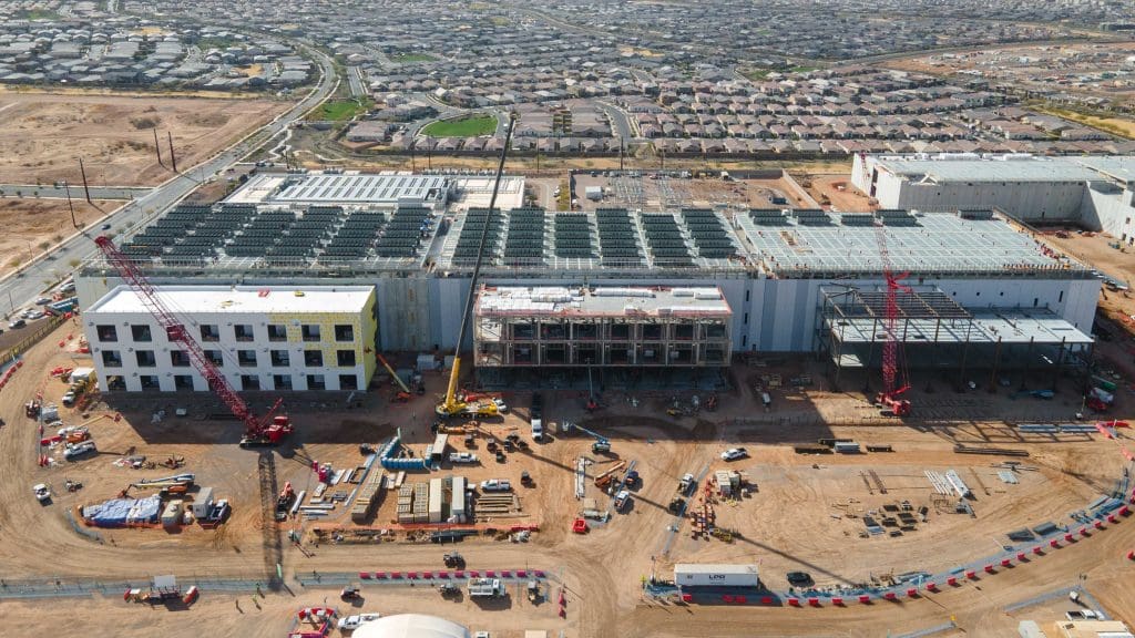 Aerial view of a large construction site with cranes, steel frames, and partially completed buildings. Workers, construction vehicles, and materials are scattered across the dirt site. Residential homes and roads are visible in the background.