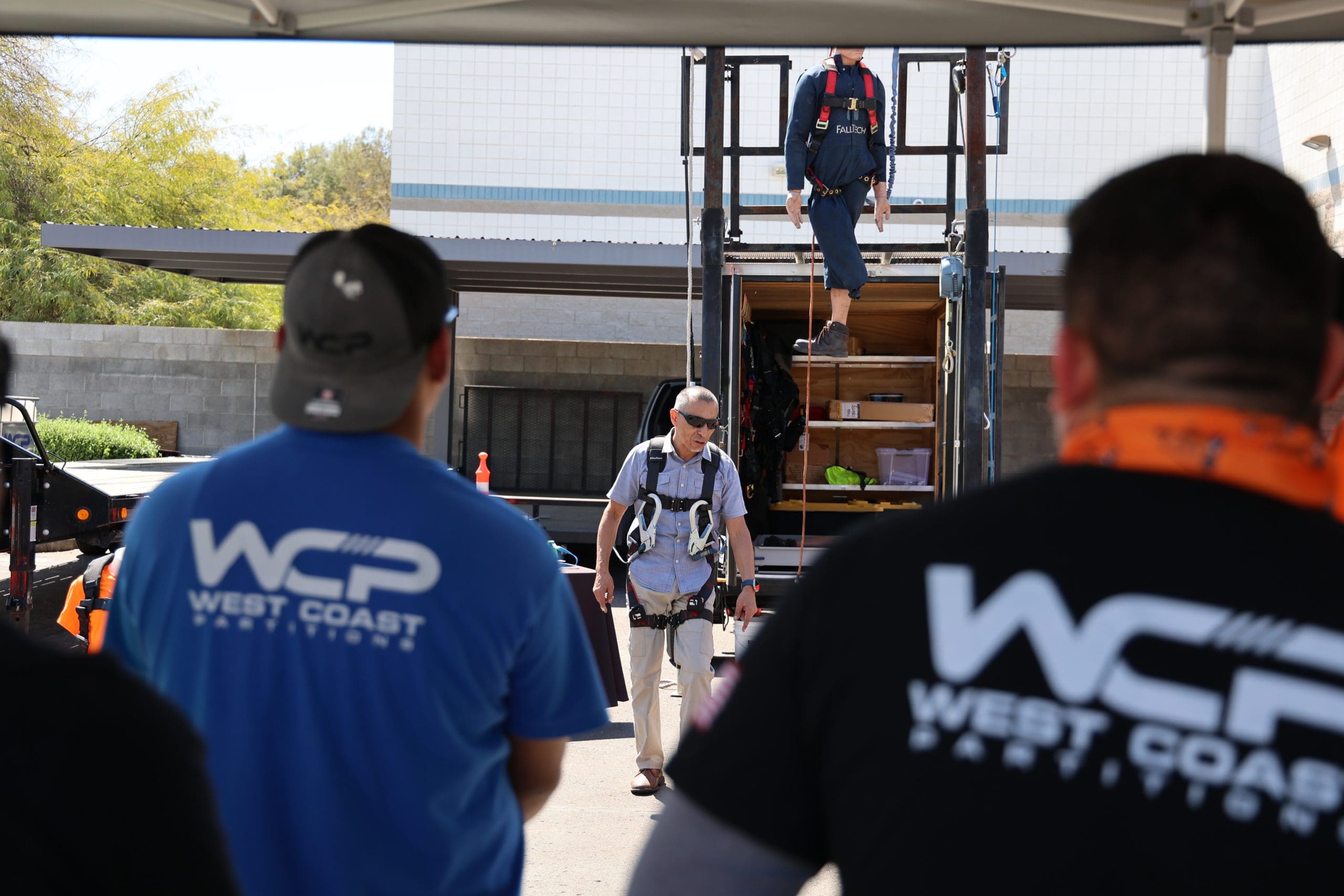 A person wearing safety gear walks away from a training structure, while two people in "WCP West Coast Partitions" shirts watch in the foreground. Another person stands at the top of the metal stairs. The scene takes place outdoors, near a building.